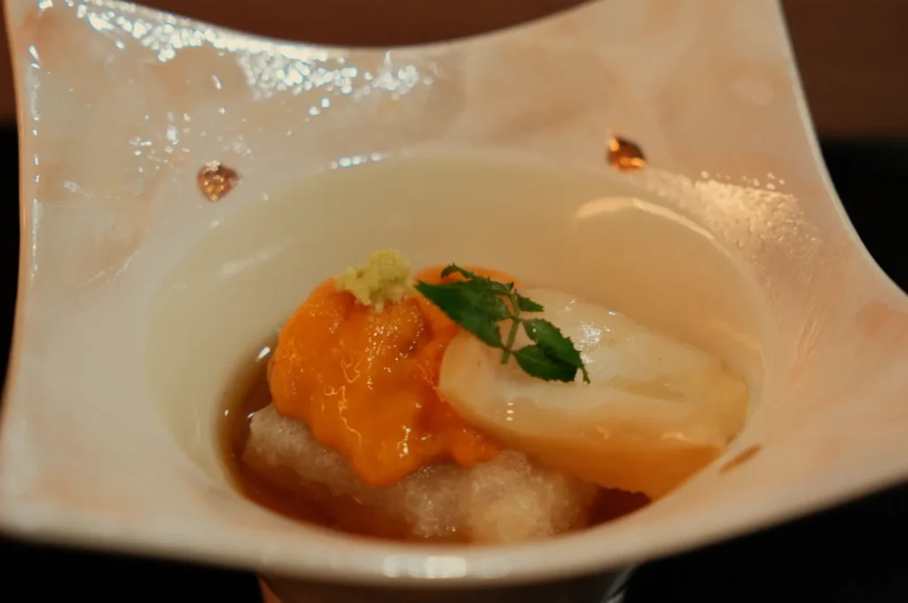 A small dish containing seafood, possibly sea urchin and a slice of scallop, garnished with a green herb and a dollop of wasabi.
