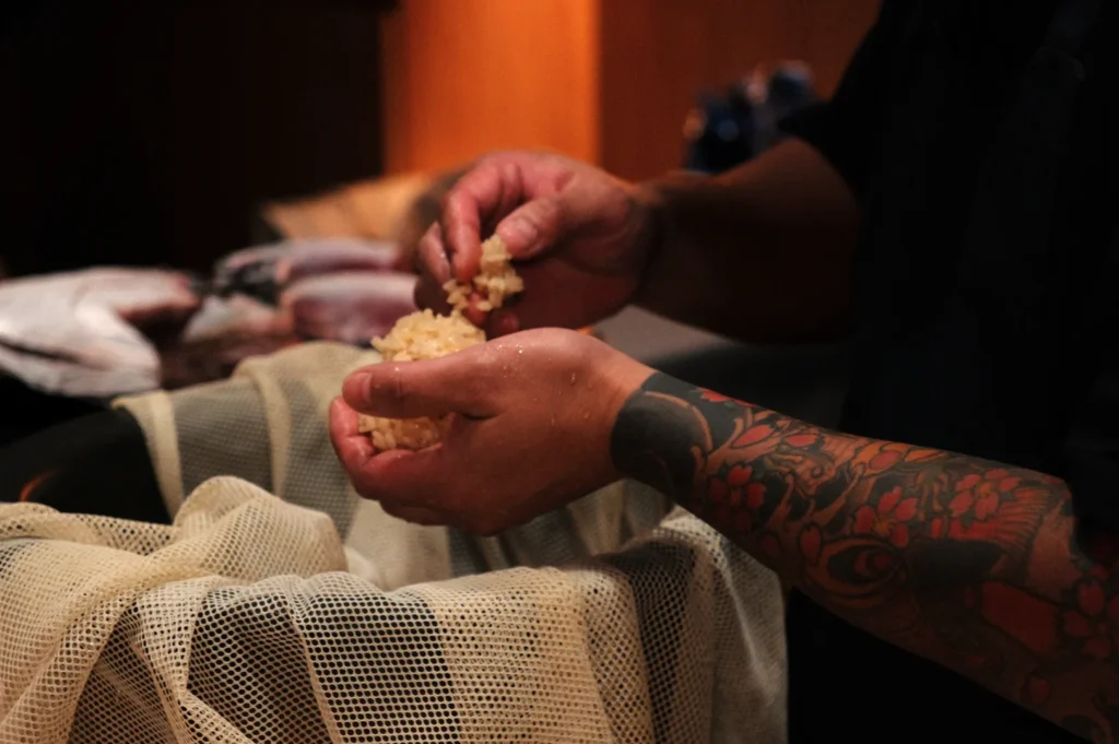 Chef Masa with tattooed arms, holding clumps of a rice substance over a mesh bag.