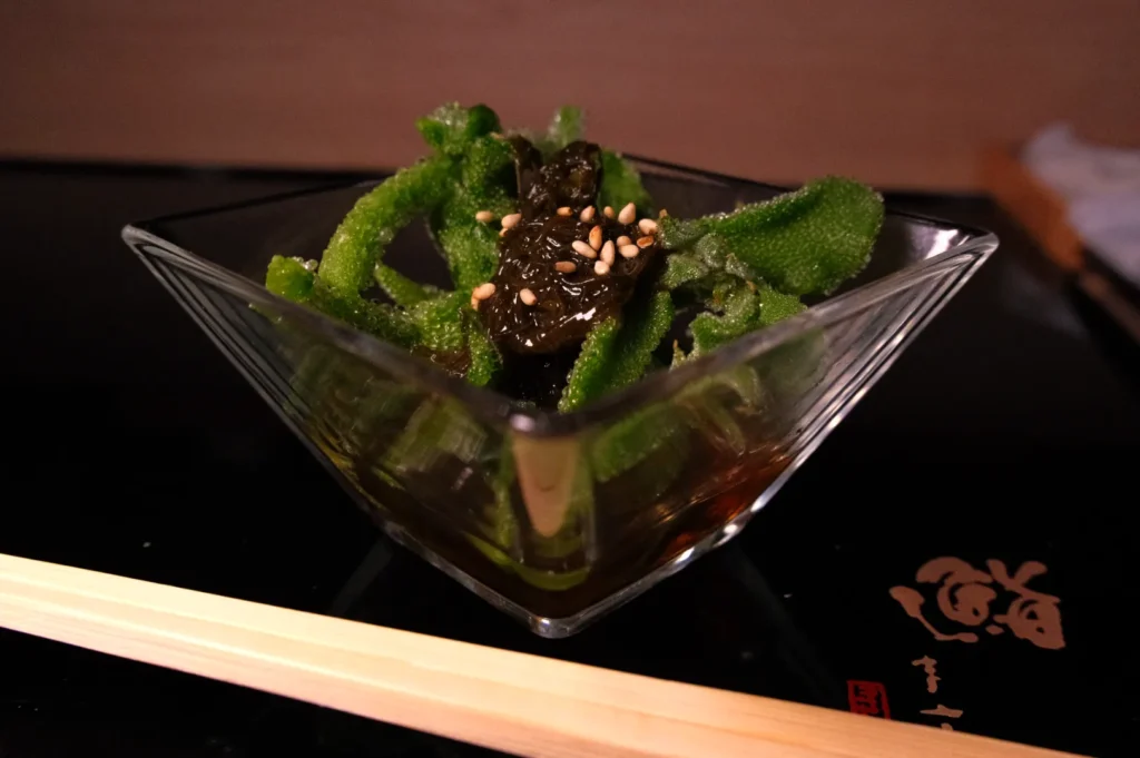 A glass bowl with green vegetables topped with sesame seeds and a dark sauce, accompanied by chopsticks on the side.