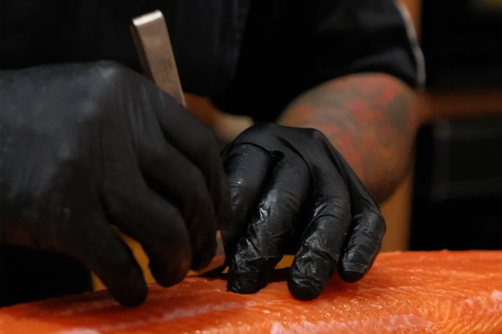 A person's hands wearing black gloves are using a tool to cut or prepare a piece of salmon.