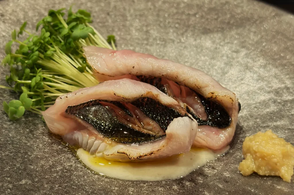 A dish featuring rolled slices of fish, garnished with microgreens and a dollop of ginger paste, served on a textured plate.