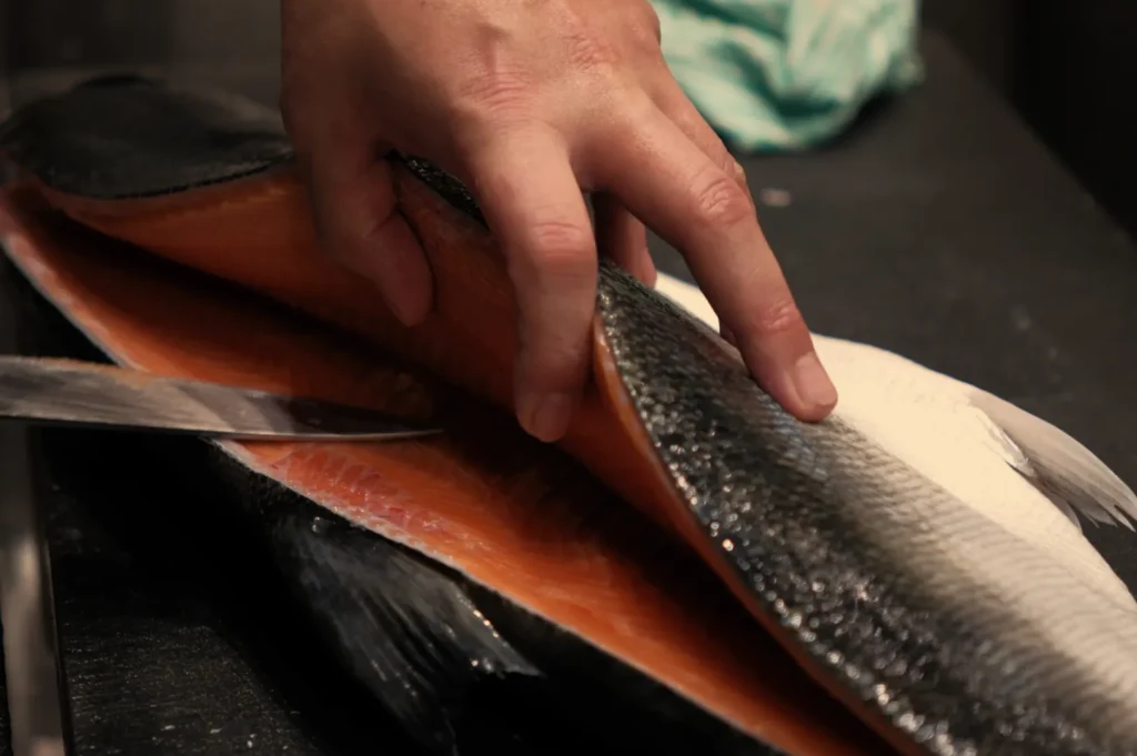A close-up of a hand holding a fillet of fish while a knife is being used to cut along the fish's body.