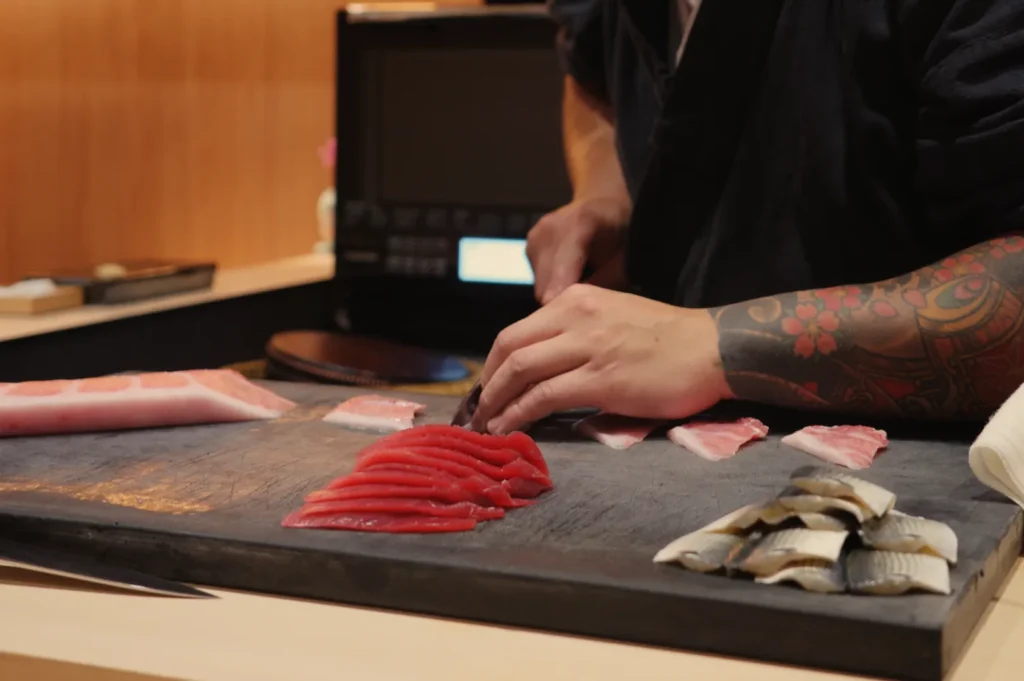 A chef with a tattoo on their arm slices fish on a cutting board, with various types of sushi displayed.