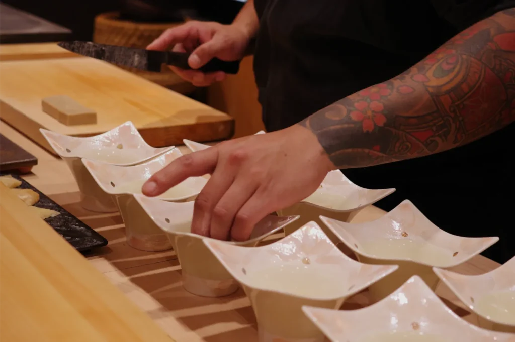 Tattooed chef carefully arranges food in white bowls on a wooden counter, with a knife nearby, creating an intimate, focused dining scene.