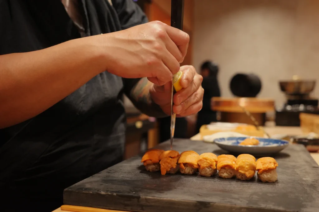 Chef Masa squeezes lemon on a knife dripping the juice on top of the uni sushi lined up on a dark board in a warmly lit kitchen.
