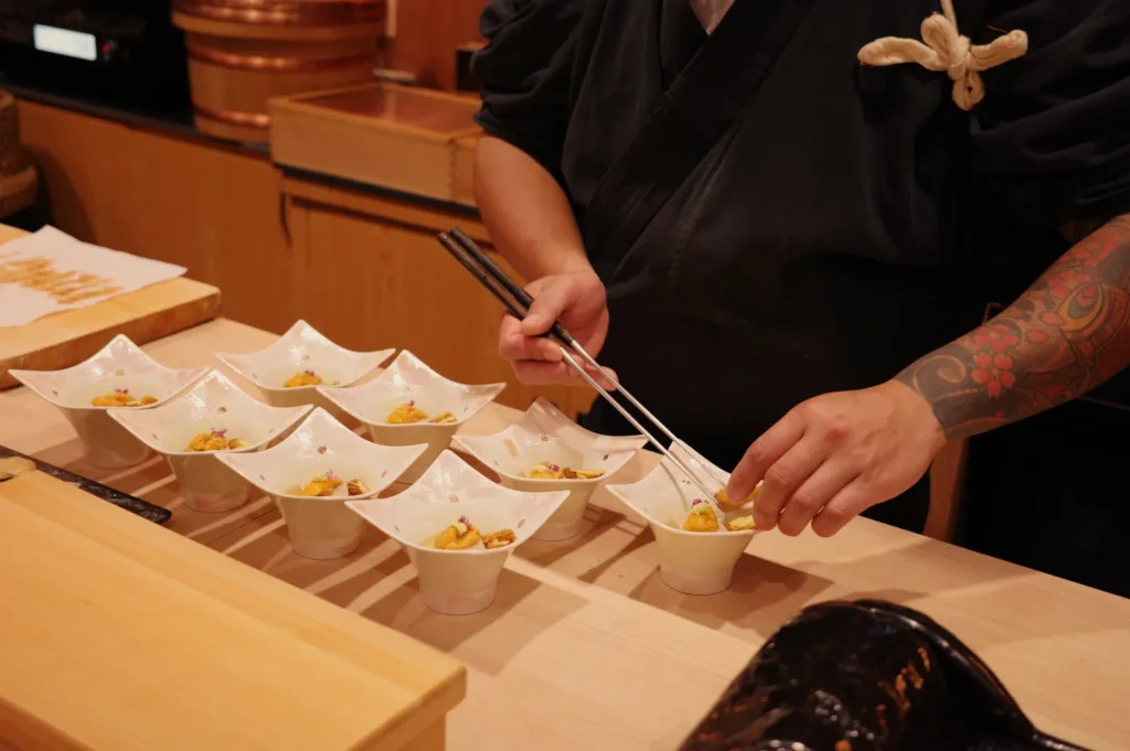 Chef Masa in traditional attire using chopsticks to arrange food in decorative bowls on a wooden countertop.