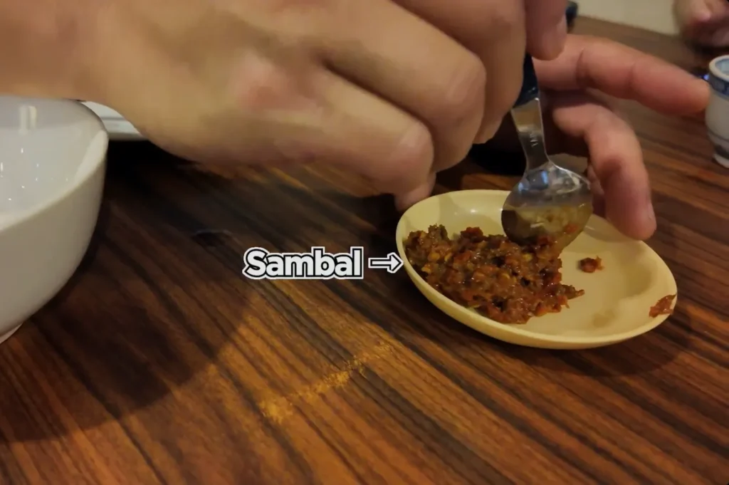 Chef Masa holding a small yellow dish and using a spoon to take a portion of sambal from the small yellow dish on a wooden table.