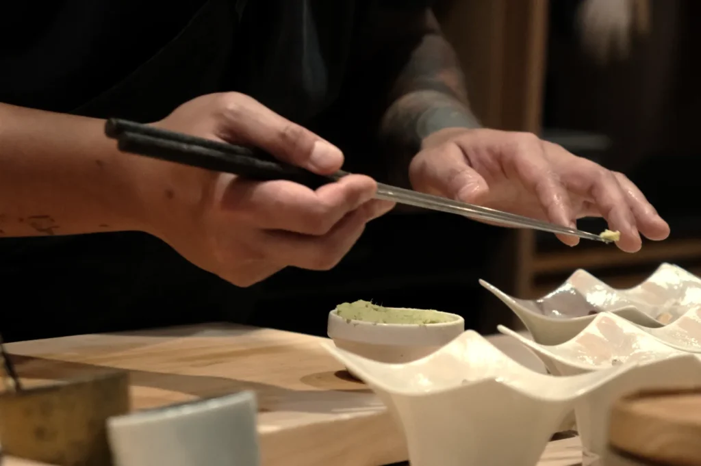 Chef Masa using chopsticks to pick up wasabi from a small dish, with several curved dishes in the foreground.