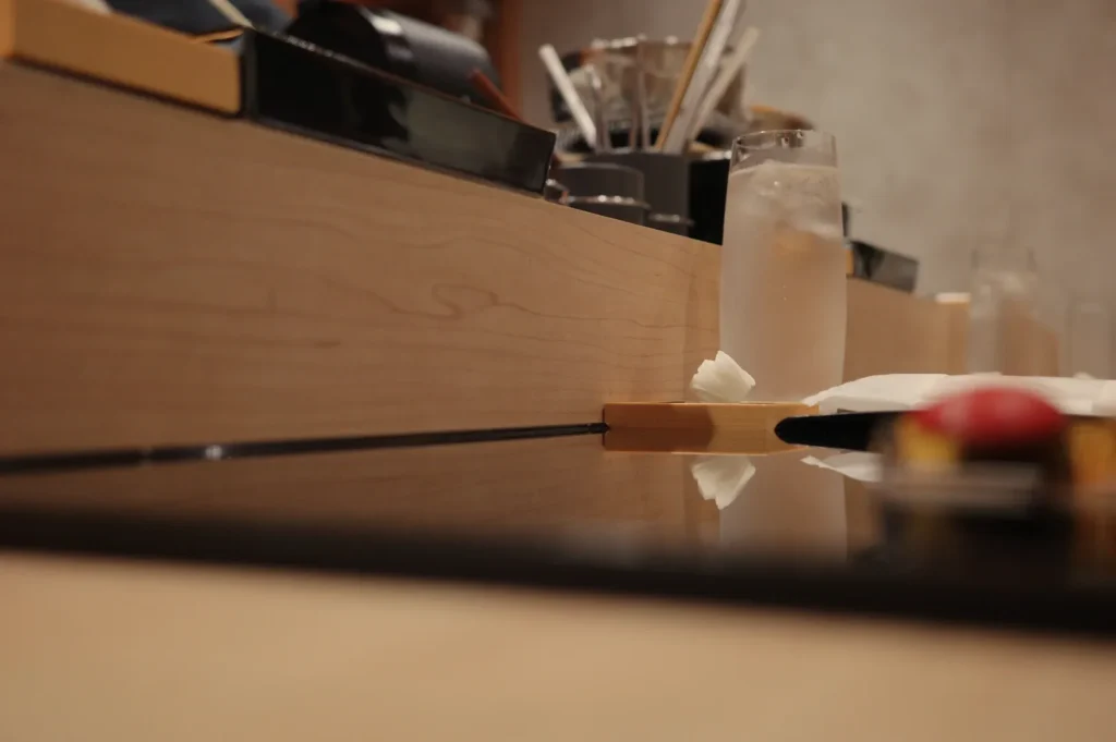Warm sushi bar scene with a wooden counter, chopsticks, iced drink, utensils, and blurred sushi in the foreground, creating a cozy mood.
