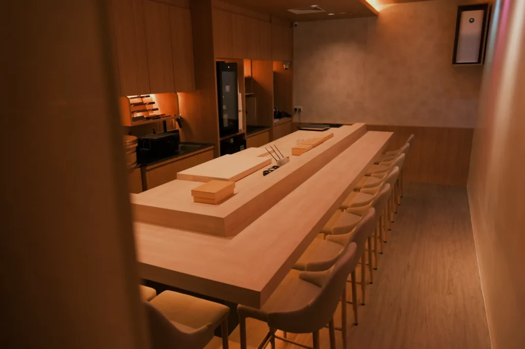 A modern, minimalist kitchen with a long wooden counter, several stools, and sleek appliances. The lighting is warm and soft, creating an inviting atmosphere.