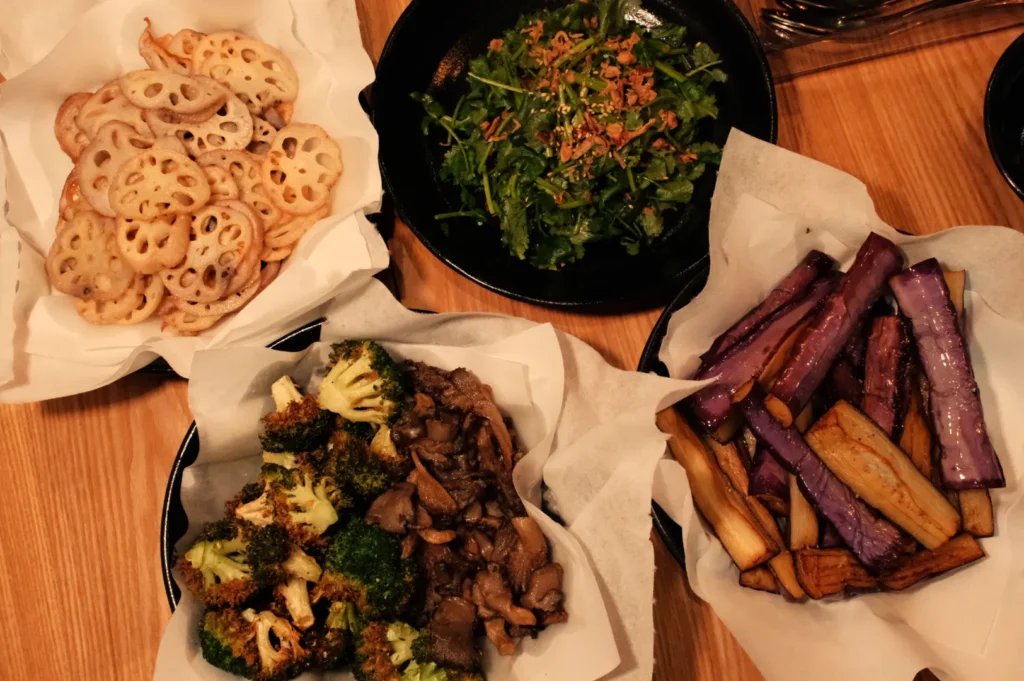 A variety of dishes including fried lotus root slices, a green salad, sautéed broccoli with mushrooms, and roasted purple and yellow vegetables, all served in bowls lined with parchment paper.