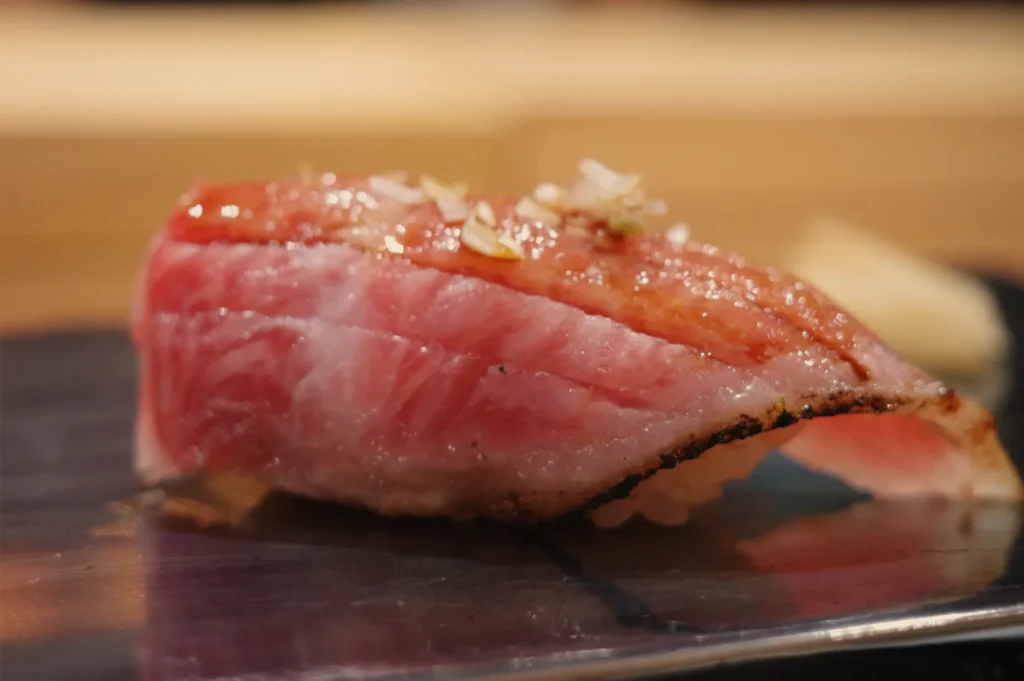Close-up of rich, marbled tuna nigiri sushi on rice, topped with sauce and garnish, resting on a dark plate; warm, appetizing mood.