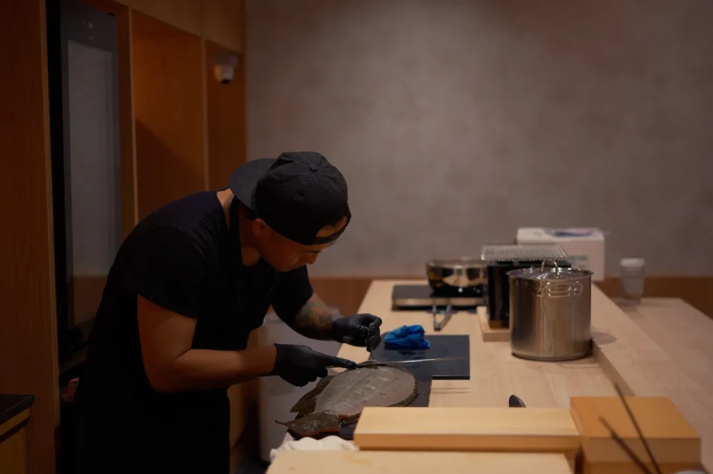 Chef Masa in black uniform and gloves carefully slices fish on a cutting board in a warm, dimly lit professional kitchen.