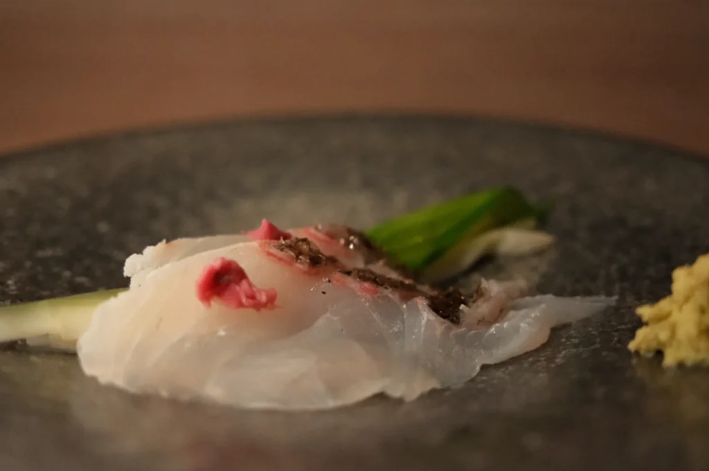 Thinly sliced white fish sashimi with red garnish, scallions, and grated ginger on a dark ceramic plate, creating an elegant, refined mood.
