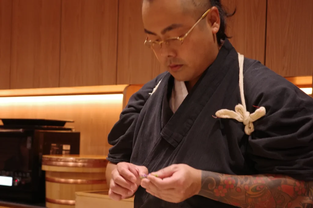 A focused sushi chef in traditional black attire carefully shapes food by hand in a warmly lit wooden restaurant kitchen, conveying calm precision and craft.
