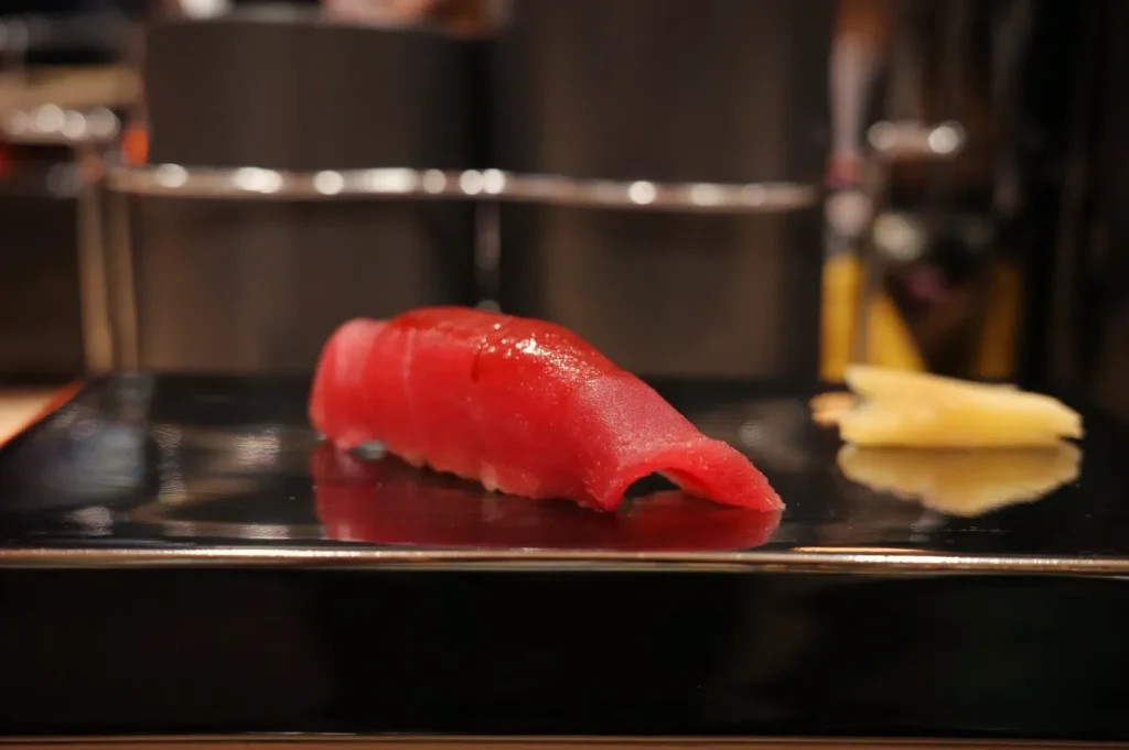 A piece of sushi with red tuna on a black plate, accompanied by a small piece of pickled ginger.