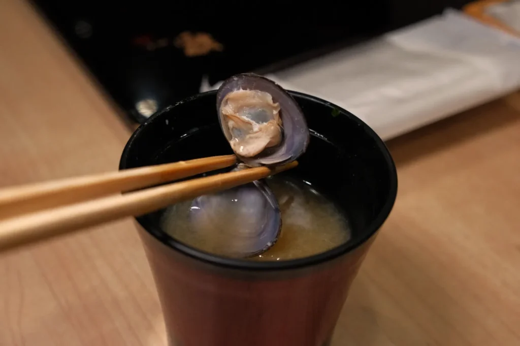 A black bowl containing clam soup, with a clam shell held by chopsticks above the bowl.