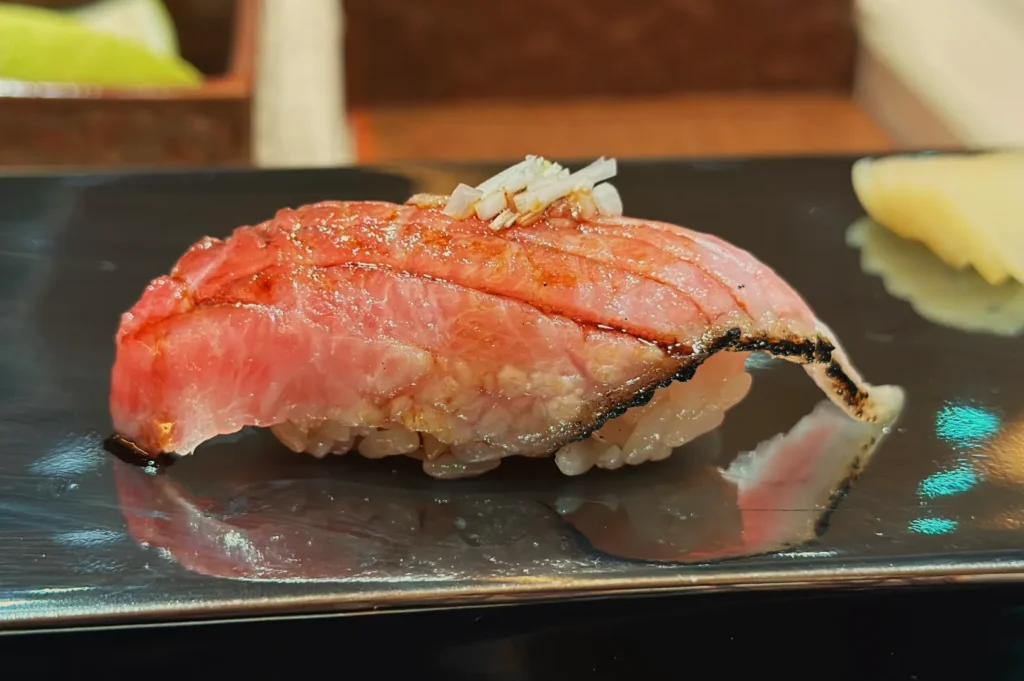 A piece of sushi featuring a slice of seared fish on top of a ball of vinegared rice, garnished with finely chopped aromatics.