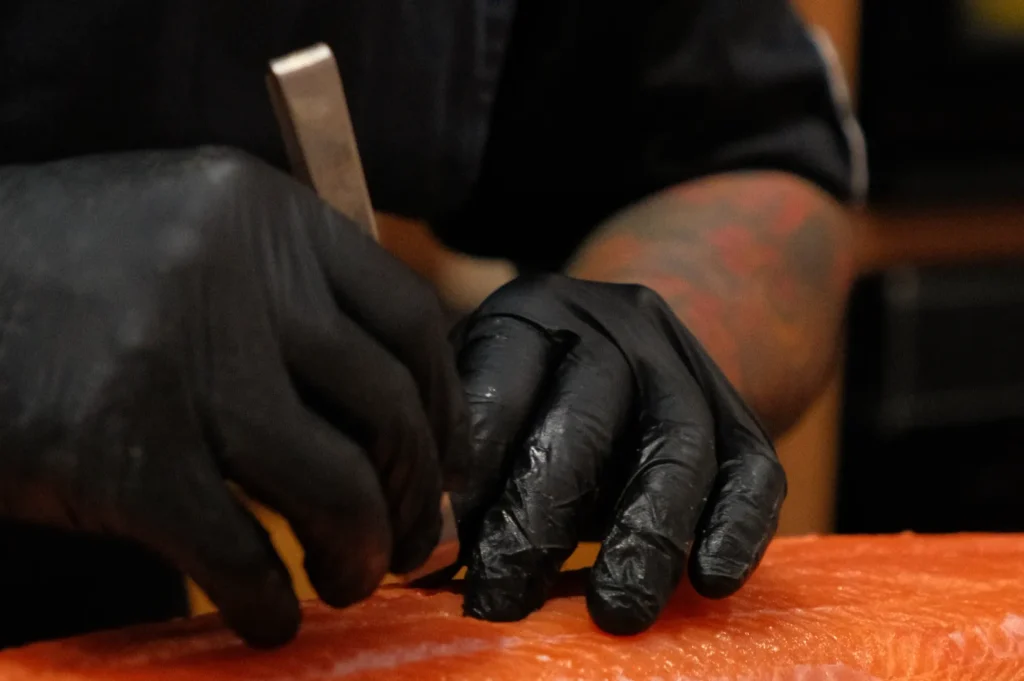 Chef Masa wearing black gloves is using a steel fish boning tweezers into a piece of salmon.