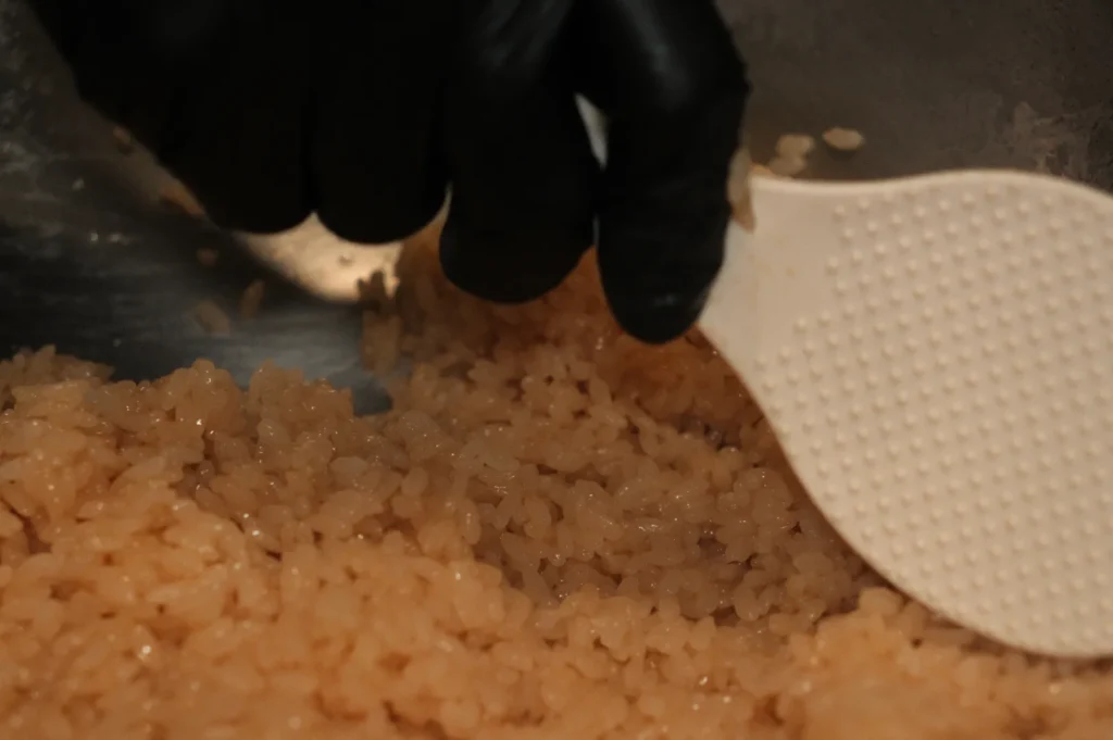 Chef Masa's hand wearing a black glove stirring cooked rice with a white spatula.