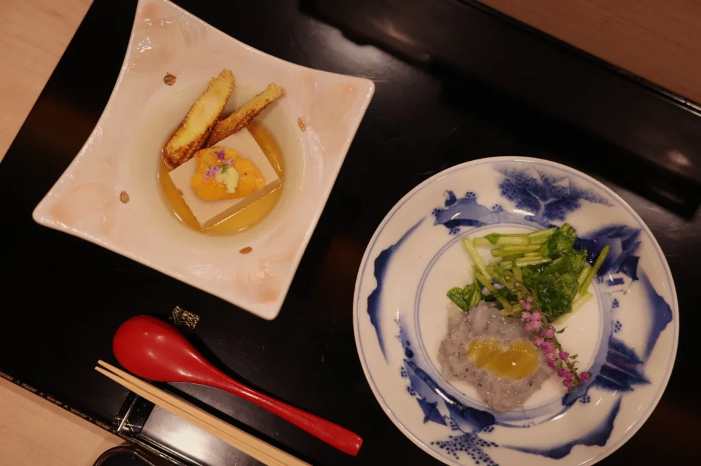 A Japanese meal on a black tray: plated fish with greens and sauce, a square dish of fries and garnish, red spoon, and chopsticks.