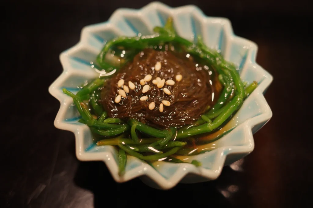 A glossy brown seaweed dish topped with sesame seeds and ringed by green vegetables, served in a pale blue-and-white star-shaped bowl on a dark table.