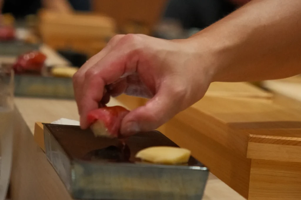 A close-up of Chef Masa’s hand placing sushi on a platter, with ginger and another piece of sushi on the side.