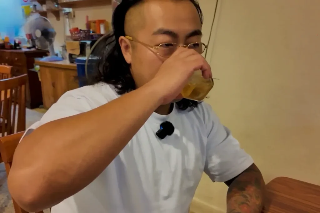 Chef Masa wearing white shirt with his hair down, sitting at a table in a restaurant, and drinking Scotch from a glass.