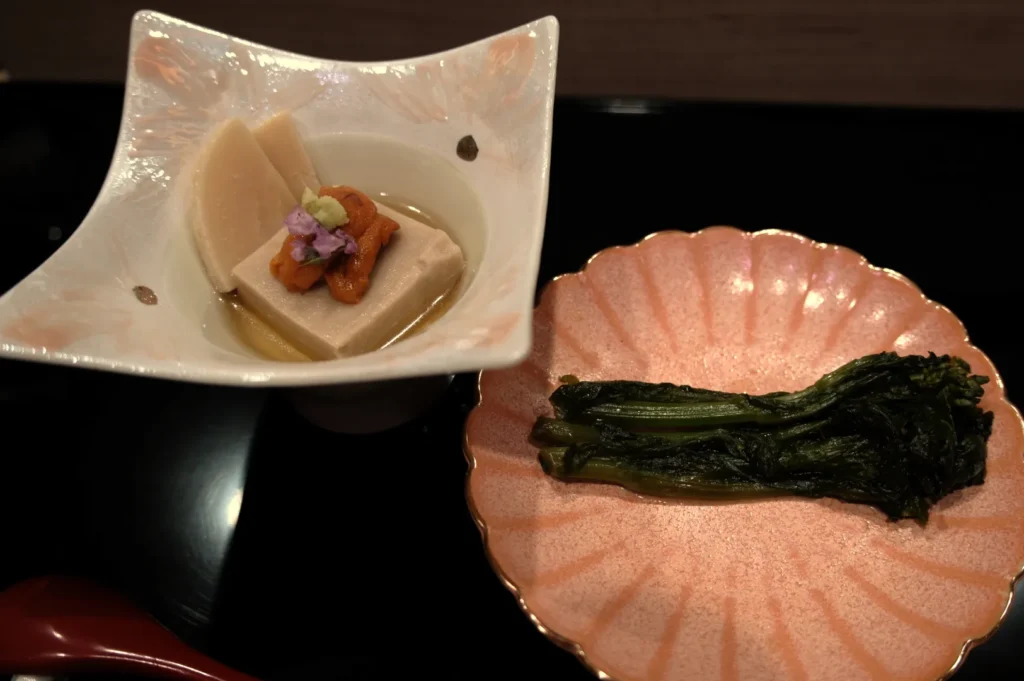 A dish consisting of tofu topped with a small amount of sauce and garnished with a flower, served alongside a dish of cooked greens.