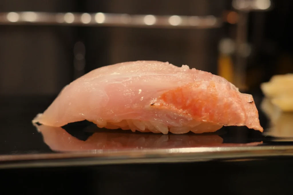A piece of sushi, specifically a slice of fish atop rice, presented on a black plate.