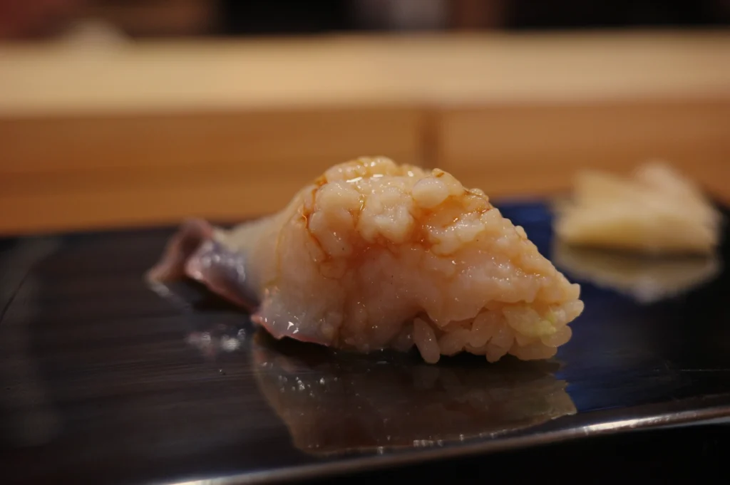 Close-up of glossy white seafood nigiri sushi on rice, served on a black plate with a softly blurred wooden counter, creating an elegant mood.