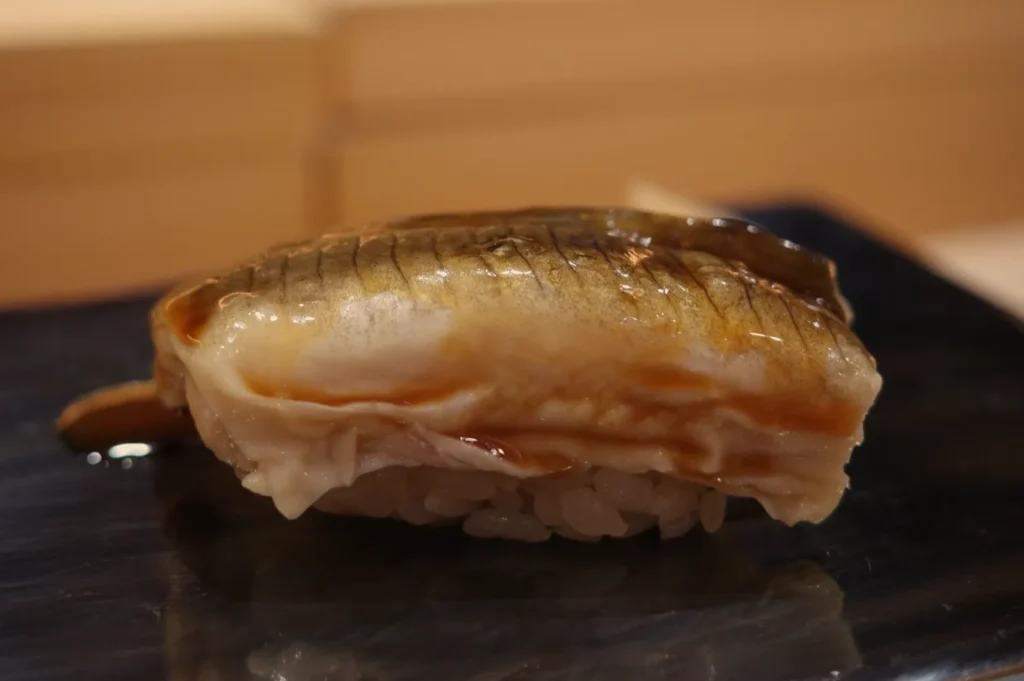 Close-up of a glossy grilled fish nigiri sushi piece with scored skin atop compact white rice, presented on a black plate in warm light.