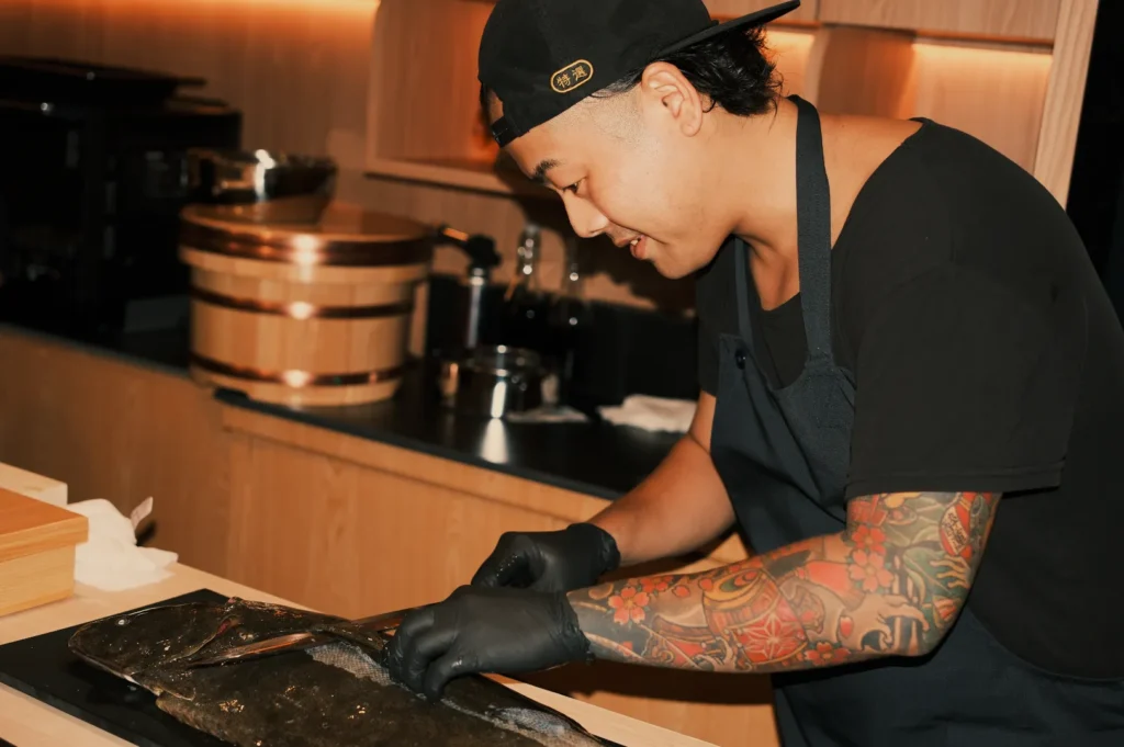 Chef in black cap, apron, and gloves prepares fish at a kitchen counter, displaying colorful tattoos and a focused, professional demeanor.