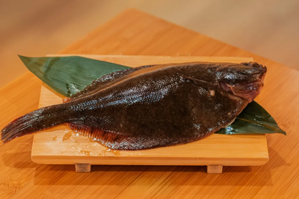 A whole dark, glossy flatfish rests on a green leaf atop a wooden cutting board, photographed against warm bamboo; the scene feels rustic and appetizing.