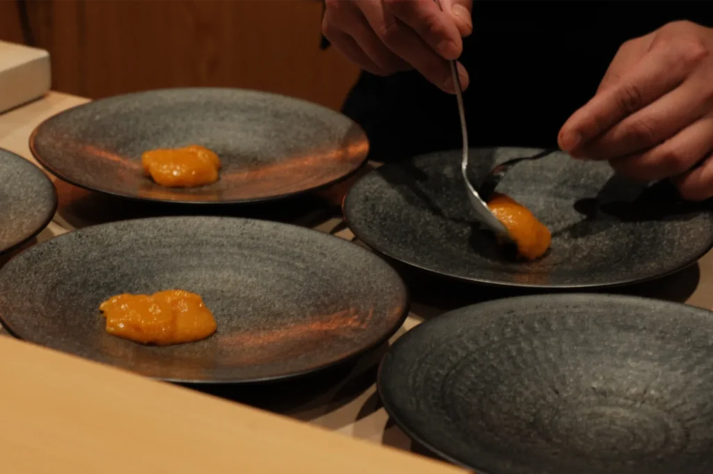 Chef Masa plating orange-colored sauce or puree onto black dishware.