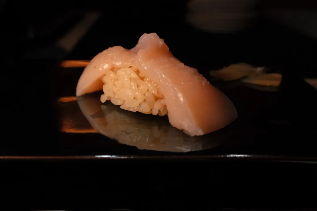 A piece of sushi featuring pale fish on top of a small ball of rice, presented on a dark plate.