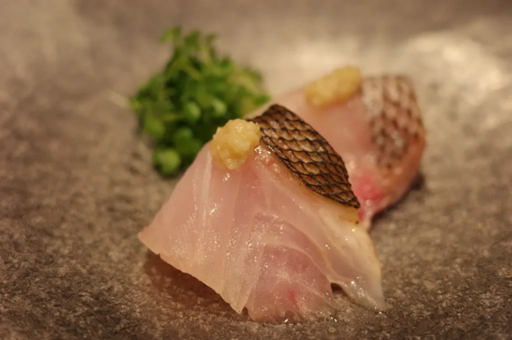 A piece of sushi featuring thinly sliced fish garnished with a small dollop of a yellow condiment, placed on a textured gray plate beside some fresh green herbs.