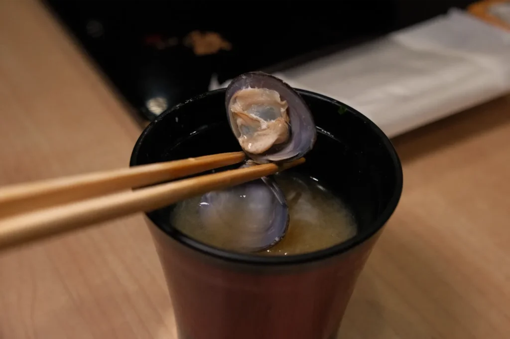 A bowl of miso soup with a piece of clam being picked up by chopsticks.