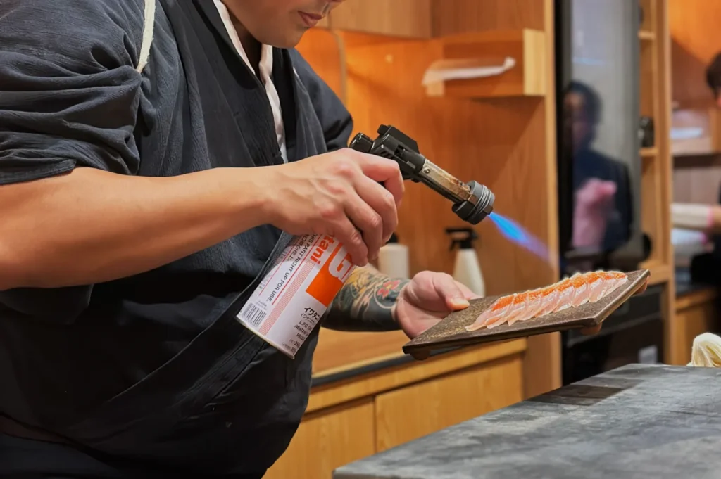 Chef Masa using a kitchen torch to caramelize or sear food on a wooden board.