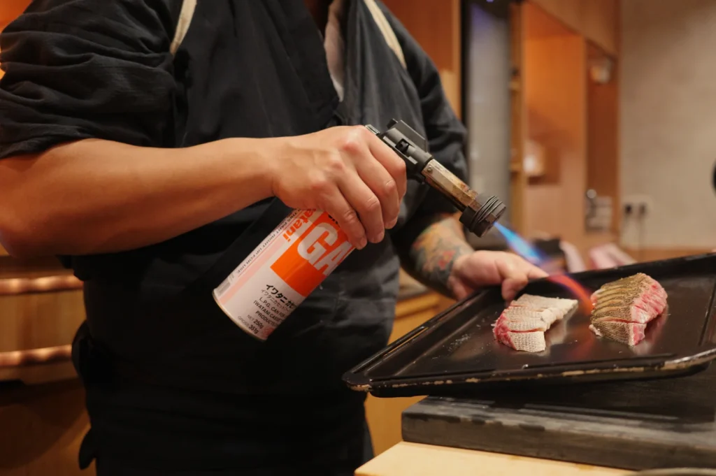 Chef Masa uses a handheld blowtorch to sear two pieces of meat on a metal tray in a warm kitchen, creating a focused, hands-on culinary scene.