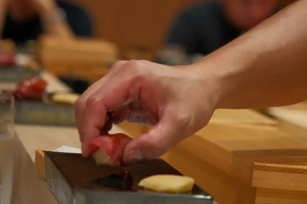 Chef Masa's hand reaching for sushi on a wooden sushi counter, with blurred people in the background.