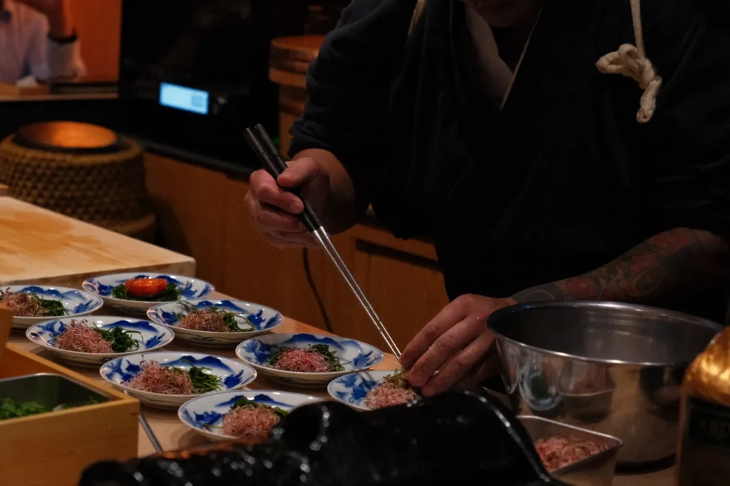 Chef Masa carefully garnishing plates of food with chopsticks in a kitchen setting.