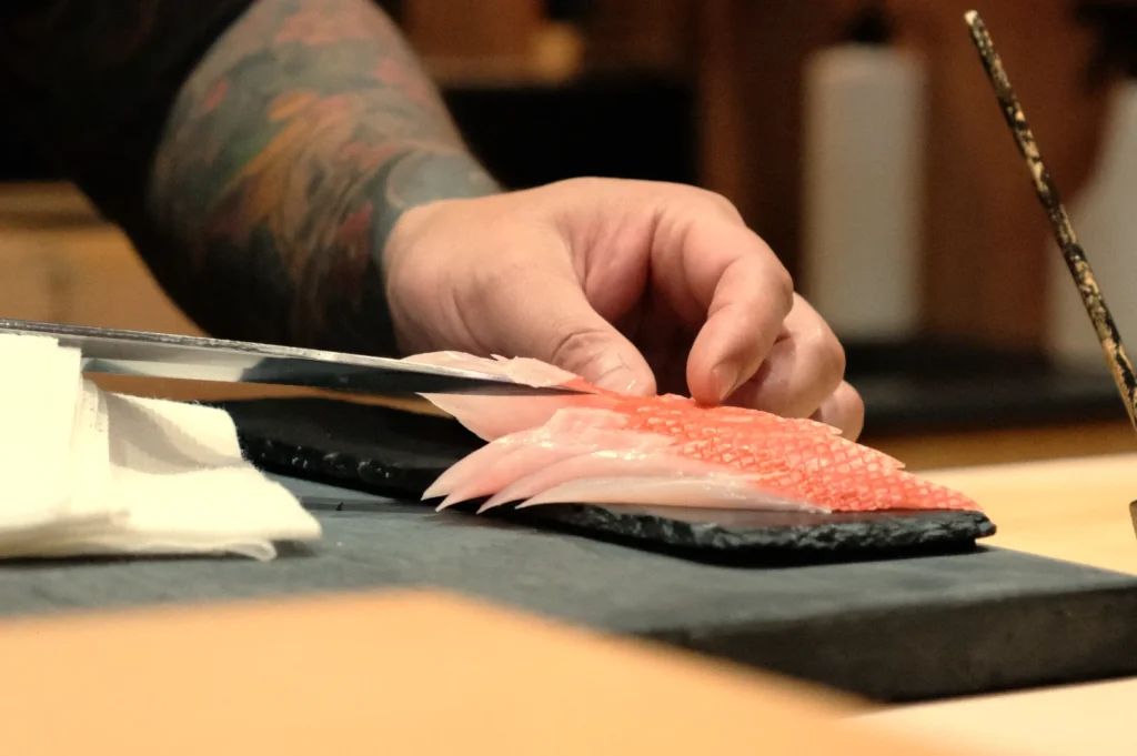 Chef Masa's hand holding a knife, slicing sushi on a black stone surface, with prepared fish slices on the surface next to the hand.