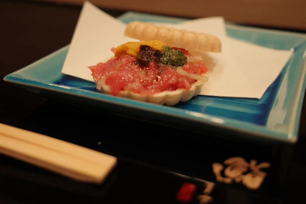 A piece of sushi presented on a blue plate, featuring a topping of raw fish and colorful garnishes, accompanied by chopsticks.