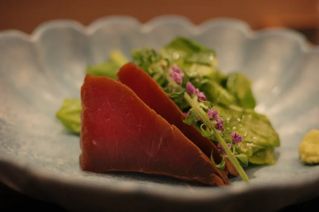 Rare seared tuna slices rest on a scalloped ceramic plate with green vegetables and delicate purple flowers, creating an elegant dish.