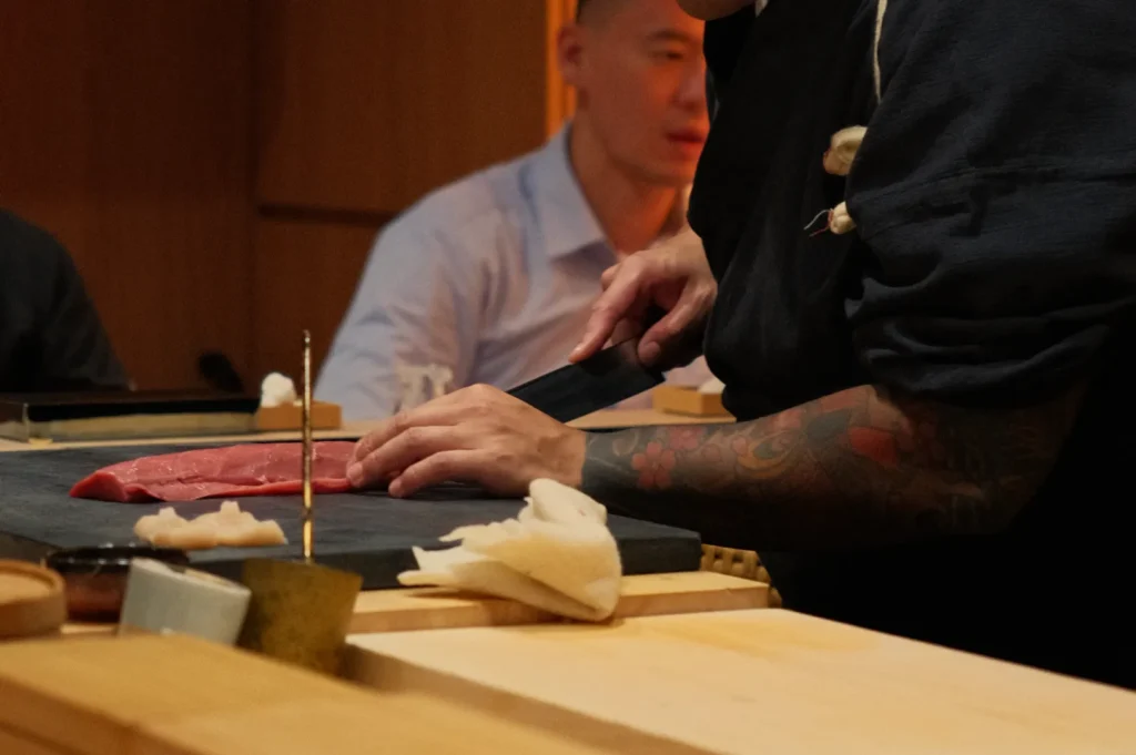 A chef preparing sushi, slicing fish on a black cutting board, with another person sitting in the background.
