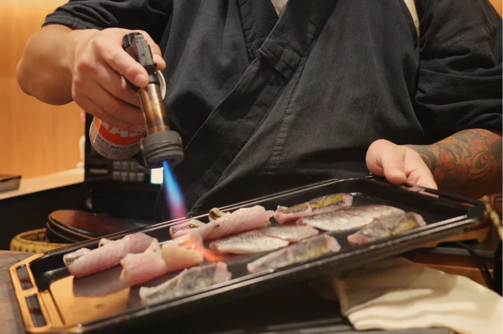 A cook in a black apron uses a blowtorch to sear sliced meat or fish on a tabletop grill, creating a dramatic blue flame in a restaurant.