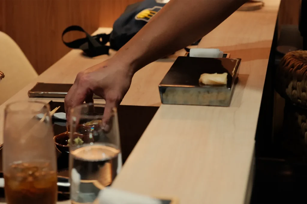 Hand reaches across a restaurant table to adjust a small glass beside water and an iced drink, with plates in warm, dim lighting.