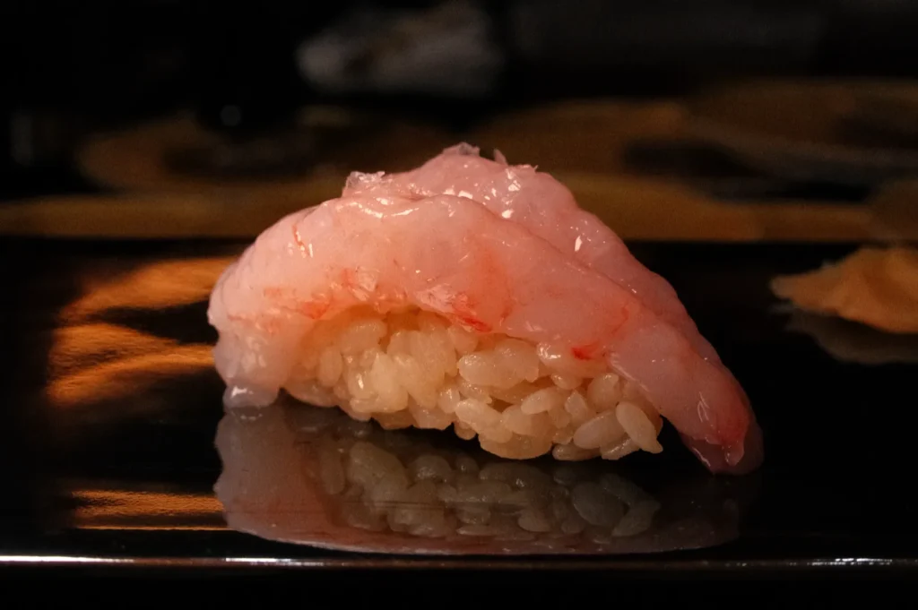 A piece of sushi featuring a slice of pink fish on top of vinegared rice, resting on a dark plate.