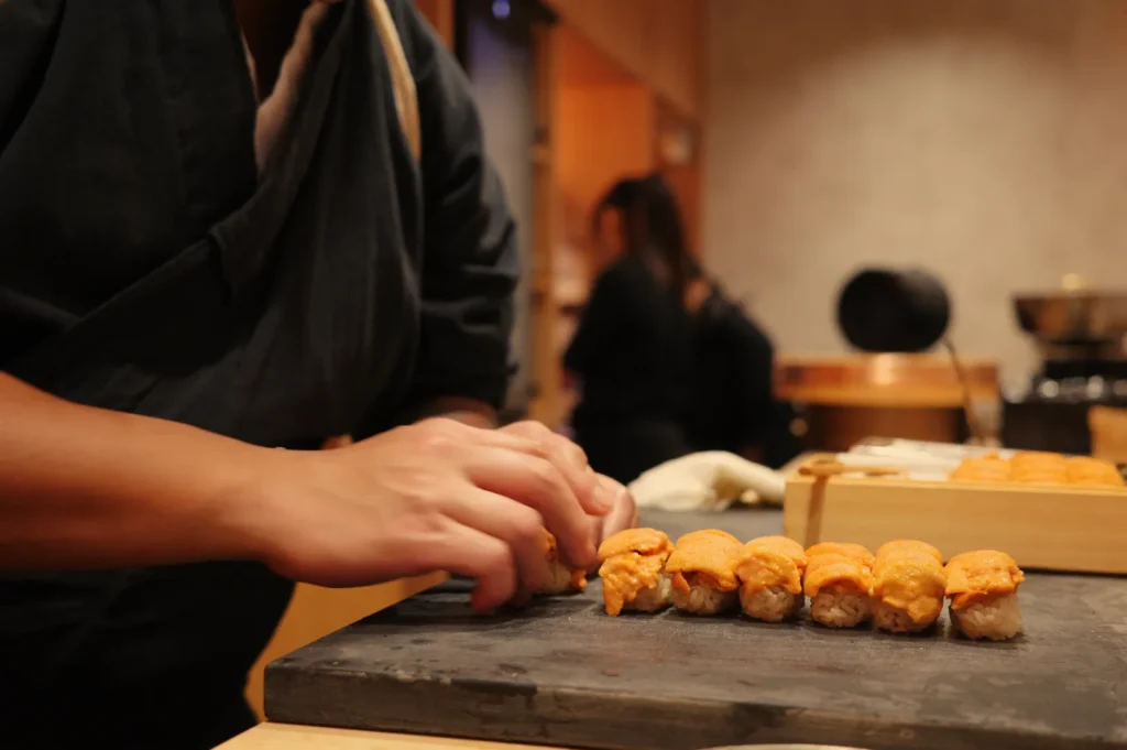 Chef Masa preparing sushi on a black stone surface, with a blurred background showing other individuals.